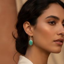 Load image into Gallery viewer, A pair of silver-toned drop earrings with oval turquoise-blue stones, displayed on a textured light blue fabric background.

