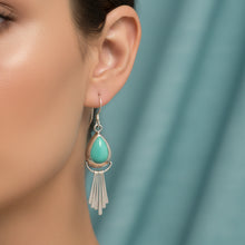 Load image into Gallery viewer, A pair of silver dangle earrings with teardrop-shaped turquoise stones and dangling fringe, displayed on a textured light blue fabric background.
