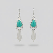 Load image into Gallery viewer, A pair of silver dangle earrings with teardrop-shaped turquoise stones and dangling fringe, displayed on a textured light blue fabric background.
