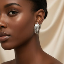 Load image into Gallery viewer, A pair of silver-toned metal earrings shaped like stylized feathers, displayed on a light  background.
